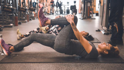 Two women in a gym setting lying on mats with one leg pulled into chest